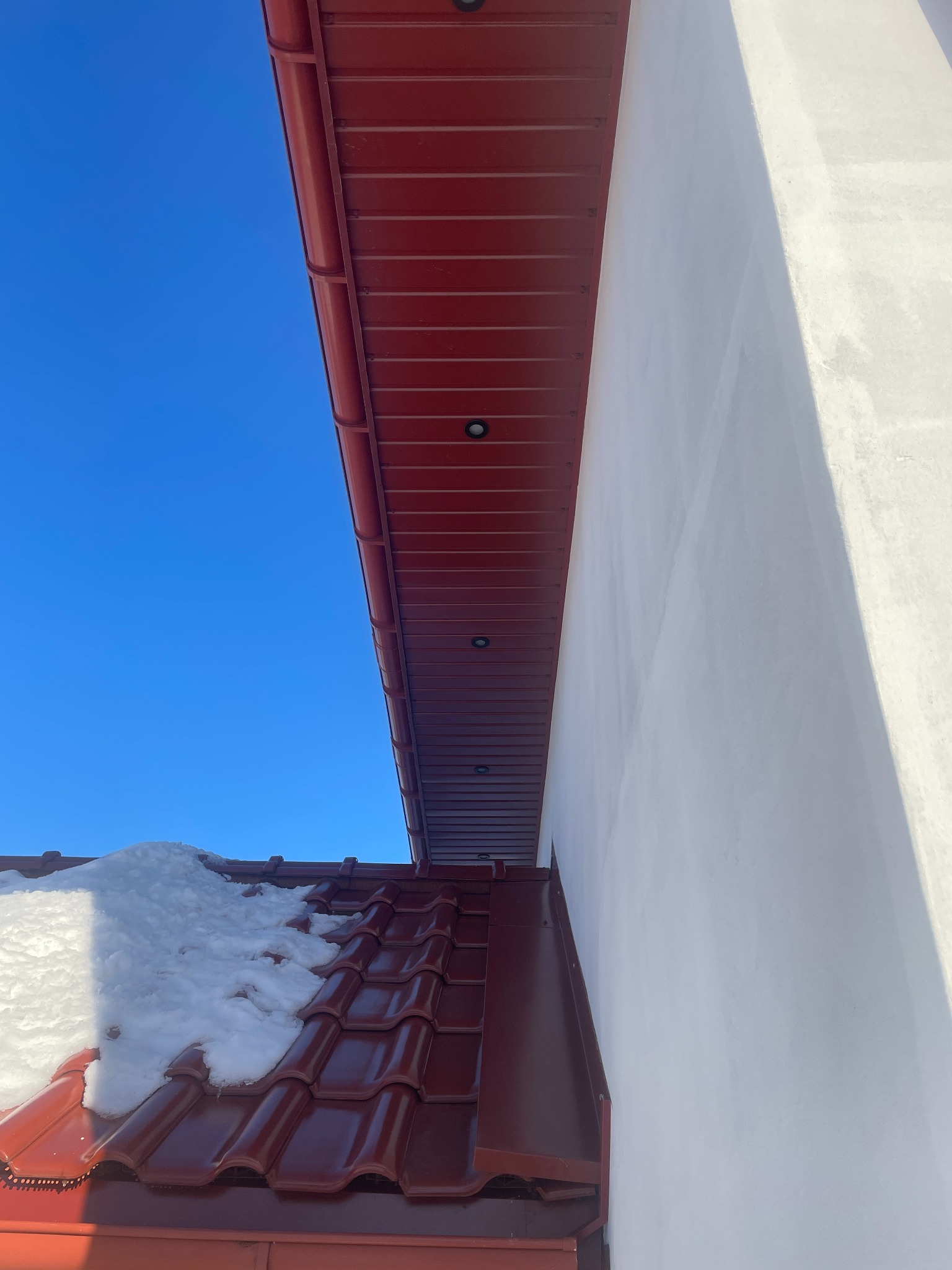 Czerwony dach z resztkami śniegu, biała ściana budynku i intensywnie błękitne niebo w słoneczny dzień. Widok z dołu, podkreślający geometrię i kontrast kolorów.