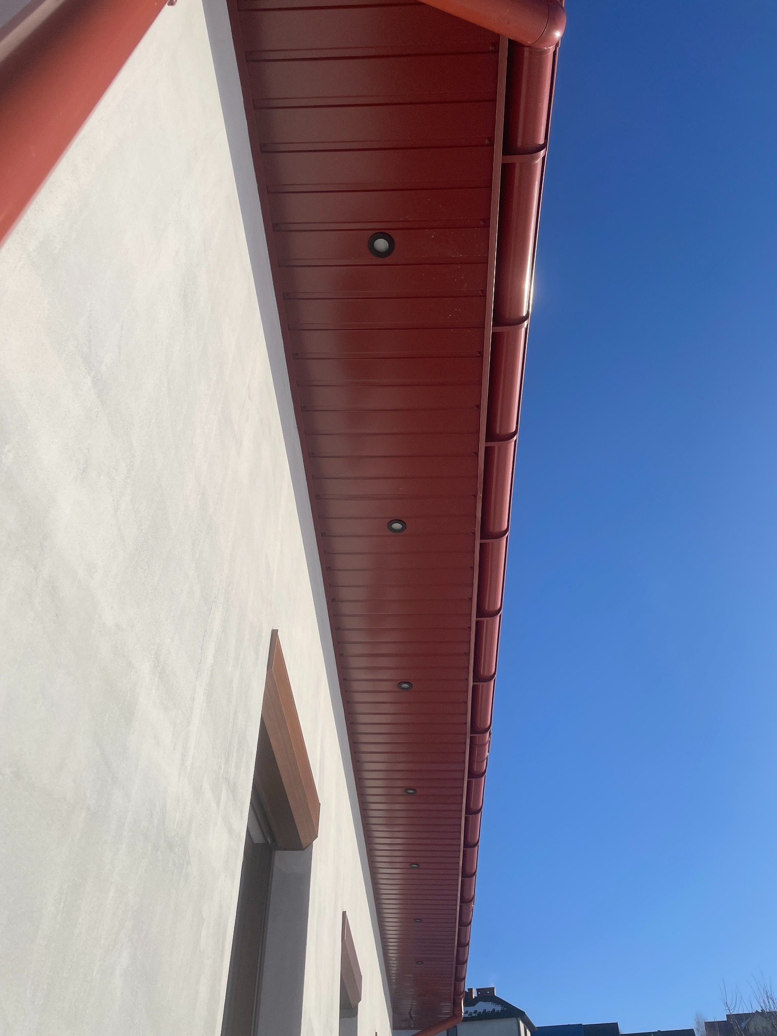 Elewacja budynku z czerwonym podbitką dachową i oświetleniem punktowym, widok z dołu na błękitne niebo. Minimalistyczny design.