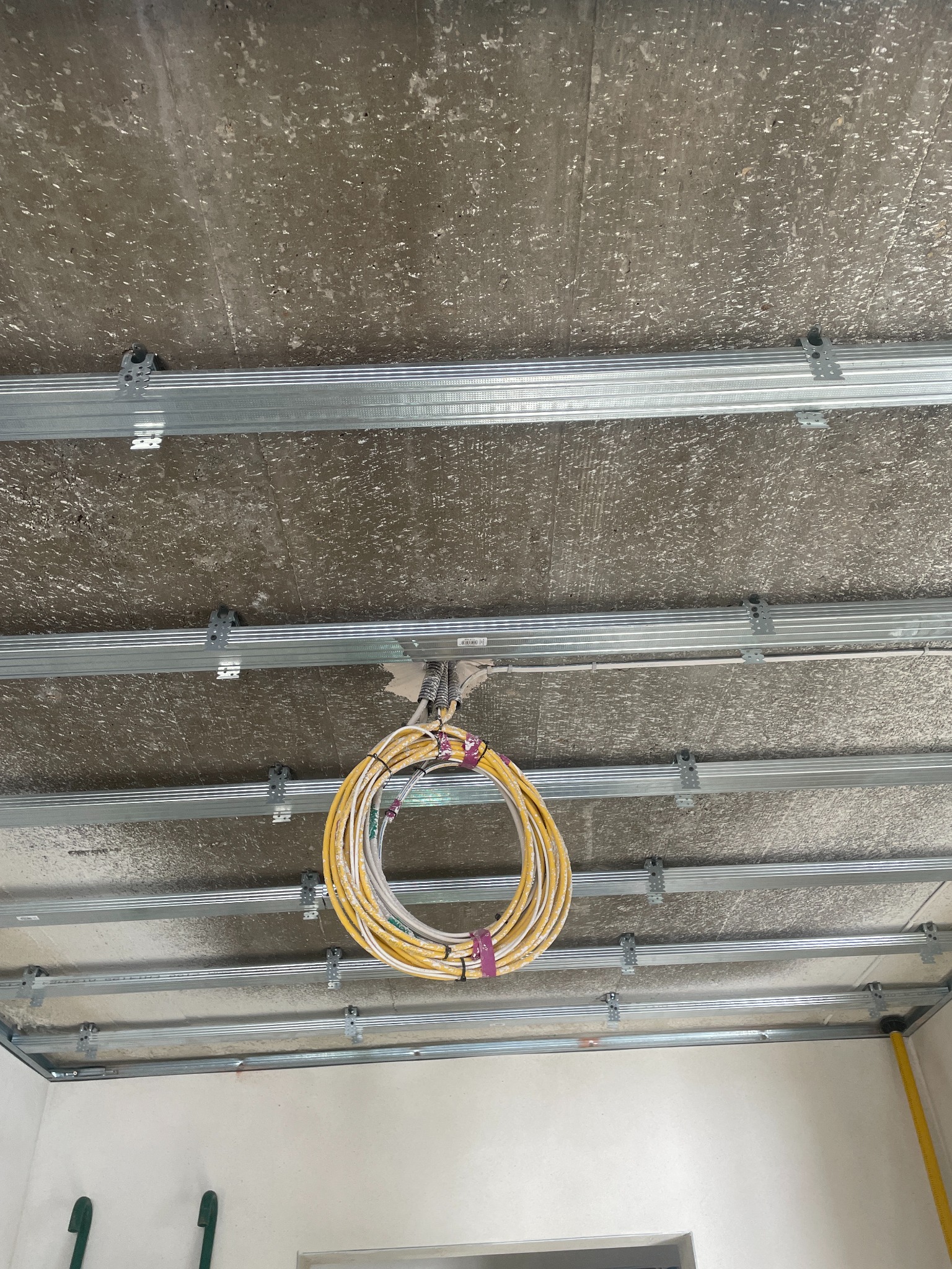 Kable elektryczne zwisające z sufitu podwieszanego z metalową konstrukcją. Widok z dołu na szare, surowe wykończenie i żółte przewody. Etap prac instalacyjnych.