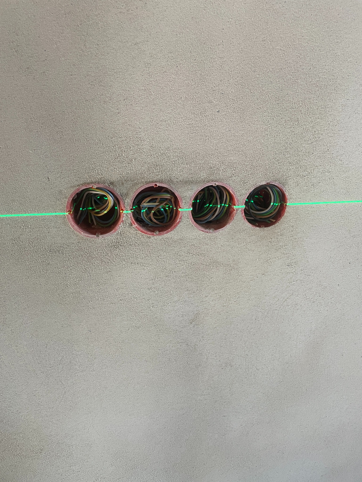 Cztery puszki elektryczne w ścianie, wyrównane laserowo, z widocznymi przewodami wewnątrz. Ściana o szarej, surowej fakturze.