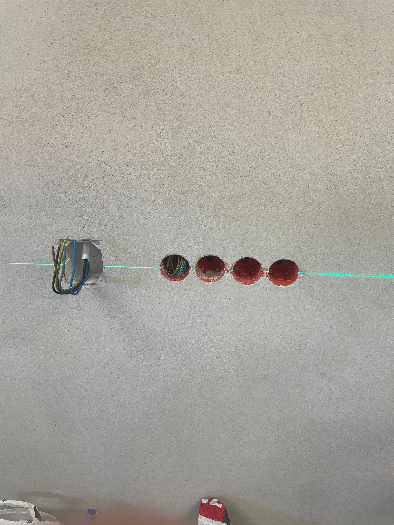 Ściana z wywierconymi otworami pod gniazdka elektryczne, widoczne przewody, poziomica laserowa wyznacza linię. Etap prac instalacyjnych.