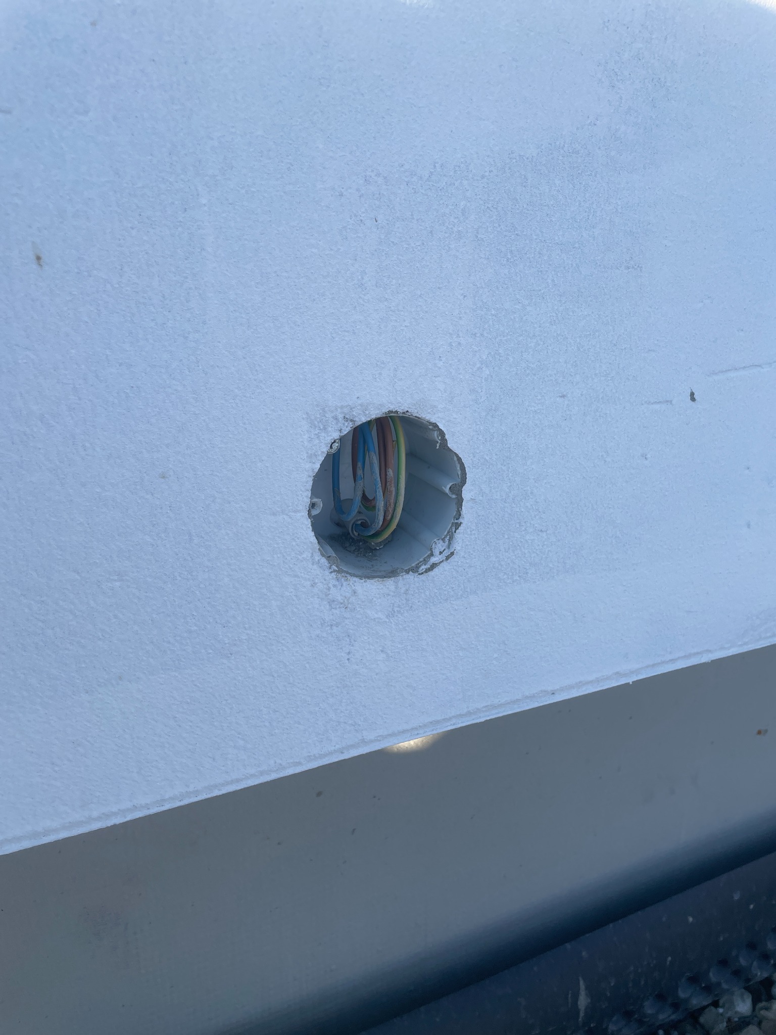 Otwór w białej ścianie z widocznymi przewodami elektrycznymi wewnątrz puszki instalacyjnej. Widok z bliska, lekki kąt.