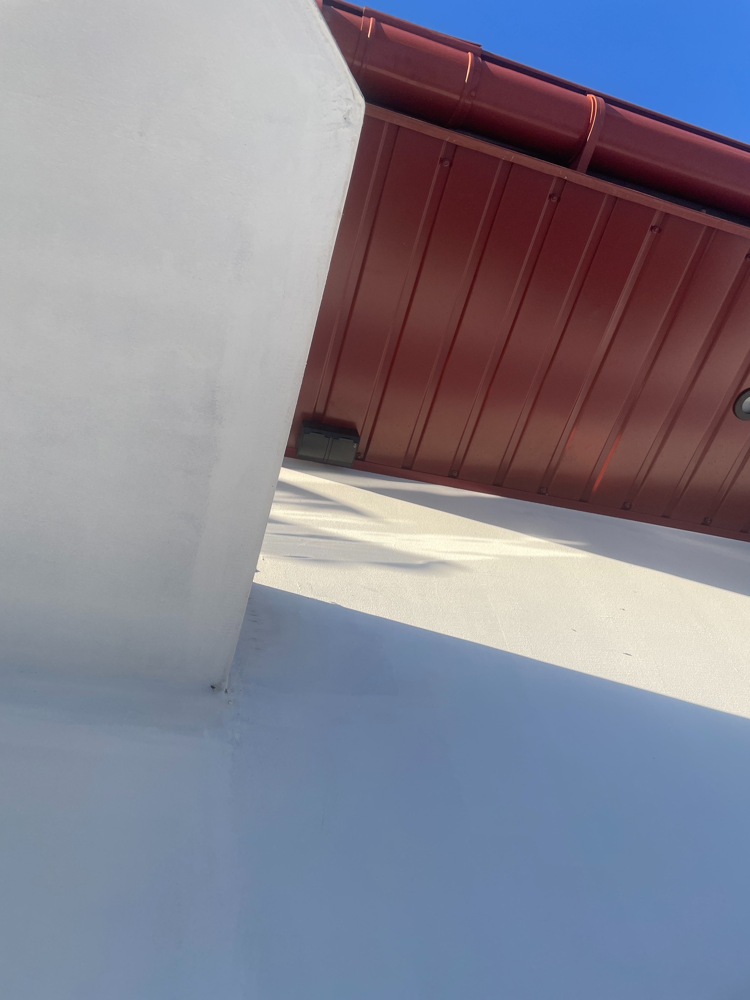 Ściana budynku po malowaniu na biało, z widocznym fragmentem dachu z blachy w kolorze ceglastym w Bącza-Kunina. Widok z dołu, oświetlenie słoneczne.