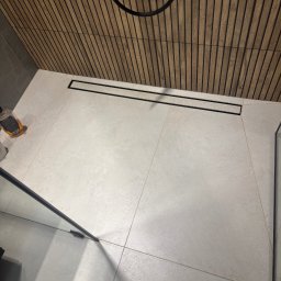 RAFBUD RAFAŁ ZIĘTKIEWICZ - Nowoczesna łazienka z jasnymi płytkami podłogowymi i drewnianą ścianą z listew. Widoczny odpływ liniowy i fragment kabiny prysznicowej.