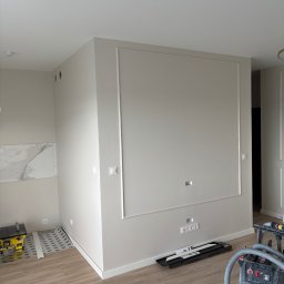 RAFBUD RAFAŁ ZIĘTKIEWICZ - Ściana z dekoracyjną ramą i gniazdkami, fragment podłogi, odkurzacz przemysłowy. Wykończenie wnętrza w neutralnej kolorystyce, widoczne narzędzia pracy.