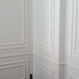 RAFBUD RAFAŁ ZIĘTKIEWICZ - Białe ściany z dekoracyjnymi listwami i sztukaterią w rogu pomieszczenia. Na podłodze karton zabezpieczający. Widoczne gniazdko elektryczne.