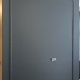 RAFBUD RAFAŁ ZIĘTKIEWICZ - Elegancka, ciemnoszara ściana z dekoracyjną ramą i listwami przypodłogowymi. Widoczne gniazdka elektryczne i narzędzia, sugerujące prace wykończeniowe.