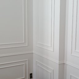 RAFBUD RAFAŁ ZIĘTKIEWICZ - Białe ściany z dekoracyjnymi listwami i sztukaterią w rogu pomieszczenia. Podłoga zabezpieczona kartonem. Widoczne gniazdko elektryczne.