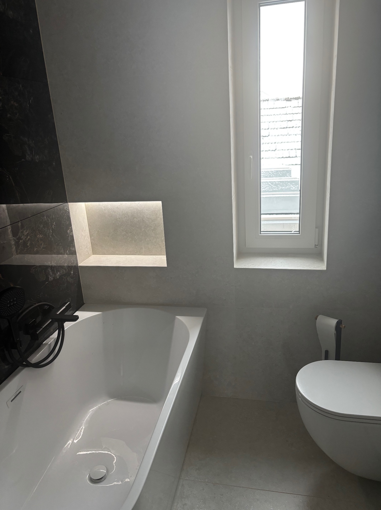 Nowoczesna łazienka z wanną, toaletą i oknem. Szare płytki imitujące kamień, czarna armatura i podświetlana wnęka nad wanną. Minimalistyczny design.