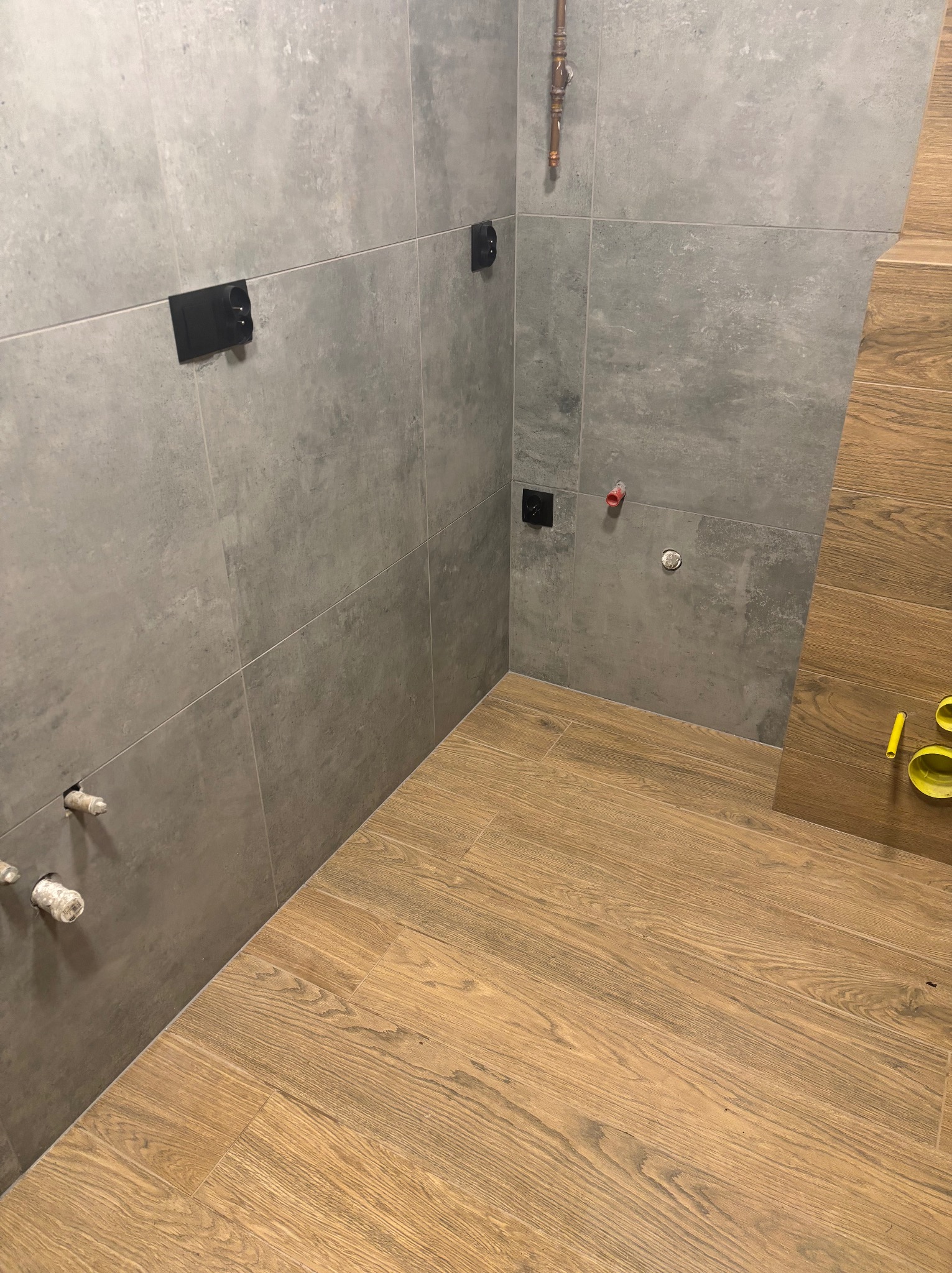 Szare płytki ścienne i drewnopodobne podłogowe w łazience, widoczne instalacje wodne i kanalizacyjne, przygotowane do montażu armatury.