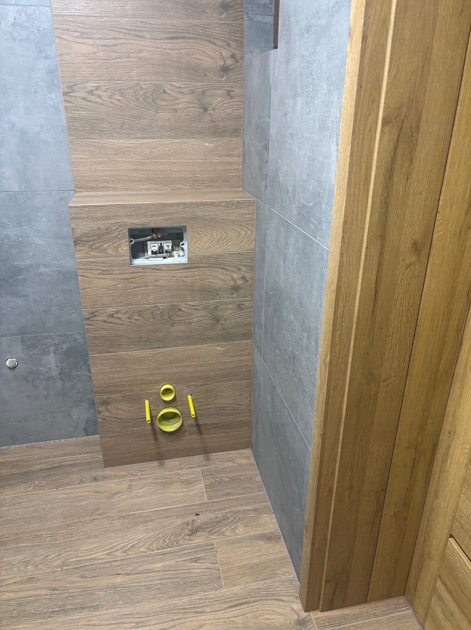 Wnętrze łazienki w trakcie wykańczania: szare płytki ścienne kontrastują z drewnopodobnymi płytkami na podłodze i zabudowie stelaża WC. Widoczne elementy instalacji.