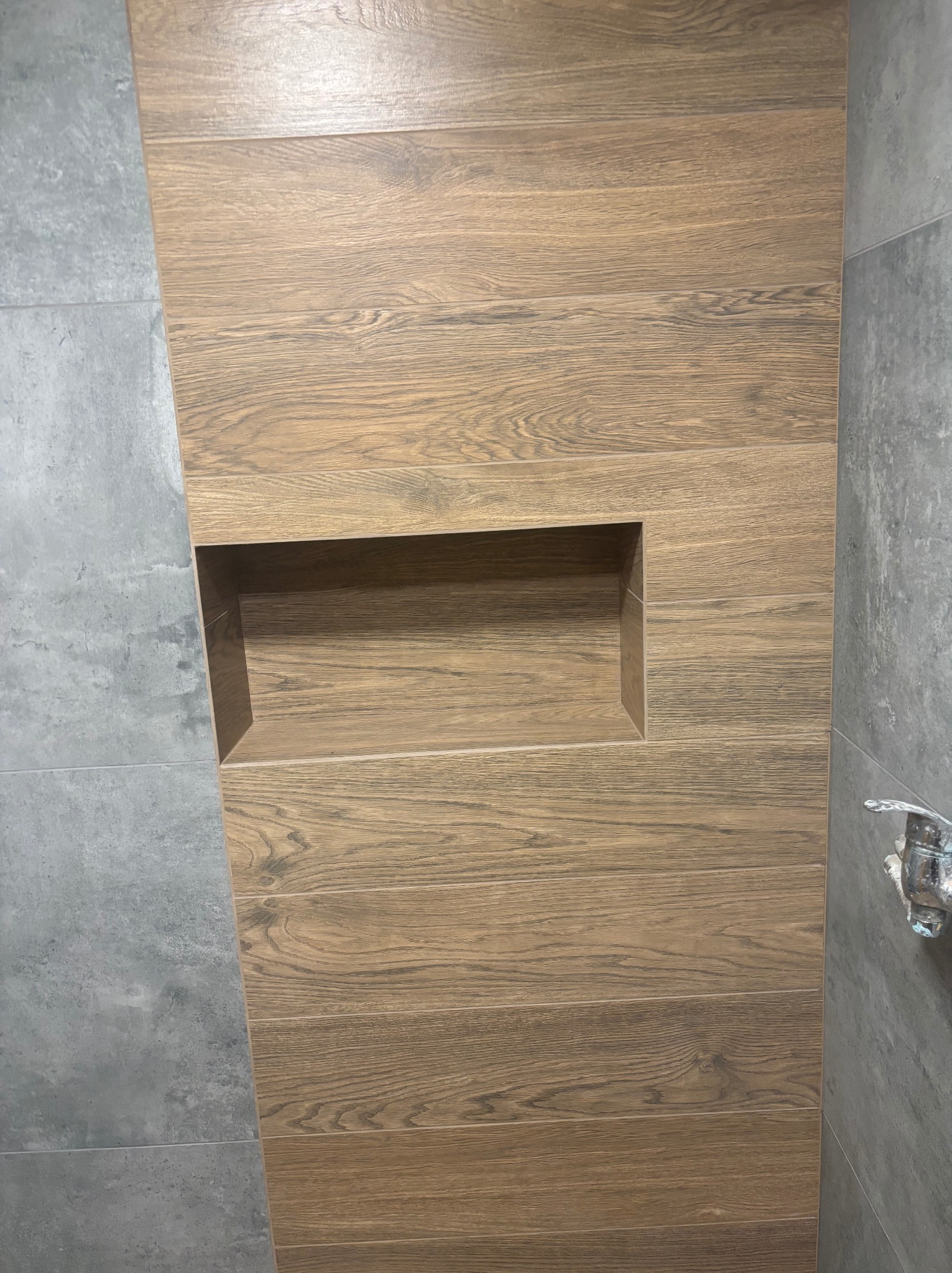 Narożna półka w kabinie prysznicowej wyłożonej płytkami imitującymi drewno, sąsiadująca z szarą, betonową ścianą. Widoczny fragment baterii.