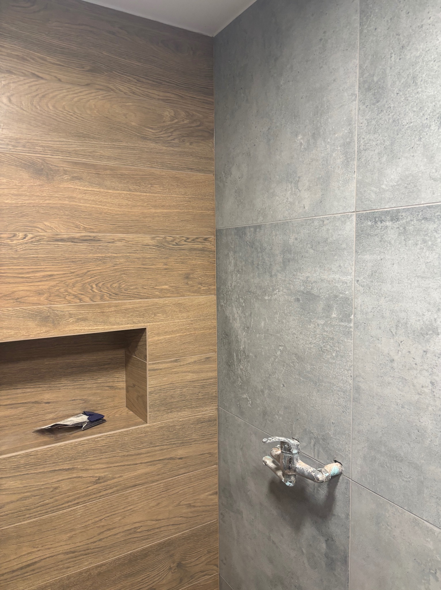 Wykończona ściana łazienki z drewnopodobnych płytek z wnęką i szarych płytek z wystającą instalacją wodną. Widoczne fugi i detale wykończenia.