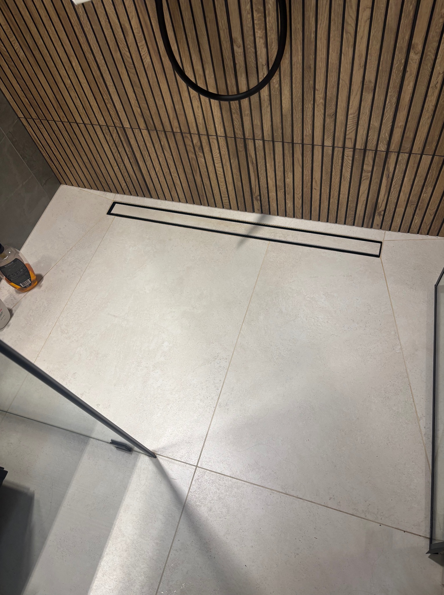 Nowoczesna łazienka z jasnymi płytkami podłogowymi i drewnianą ścianą z listew. Widoczny odpływ liniowy i fragment kabiny prysznicowej.