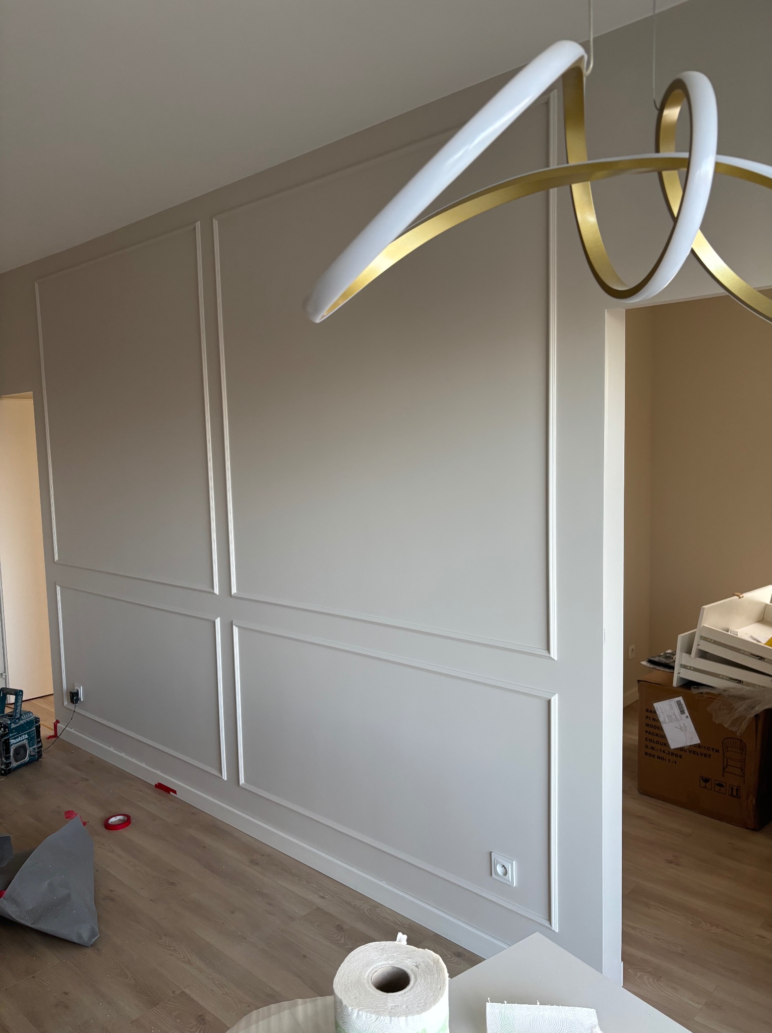 Ściana z dekoracyjnymi listwami w jasnym odcieniu, oświetlona nowoczesną lampą. Widoczne narzędzia i materiały wykończeniowe. Detal wykończenia wnętrza.