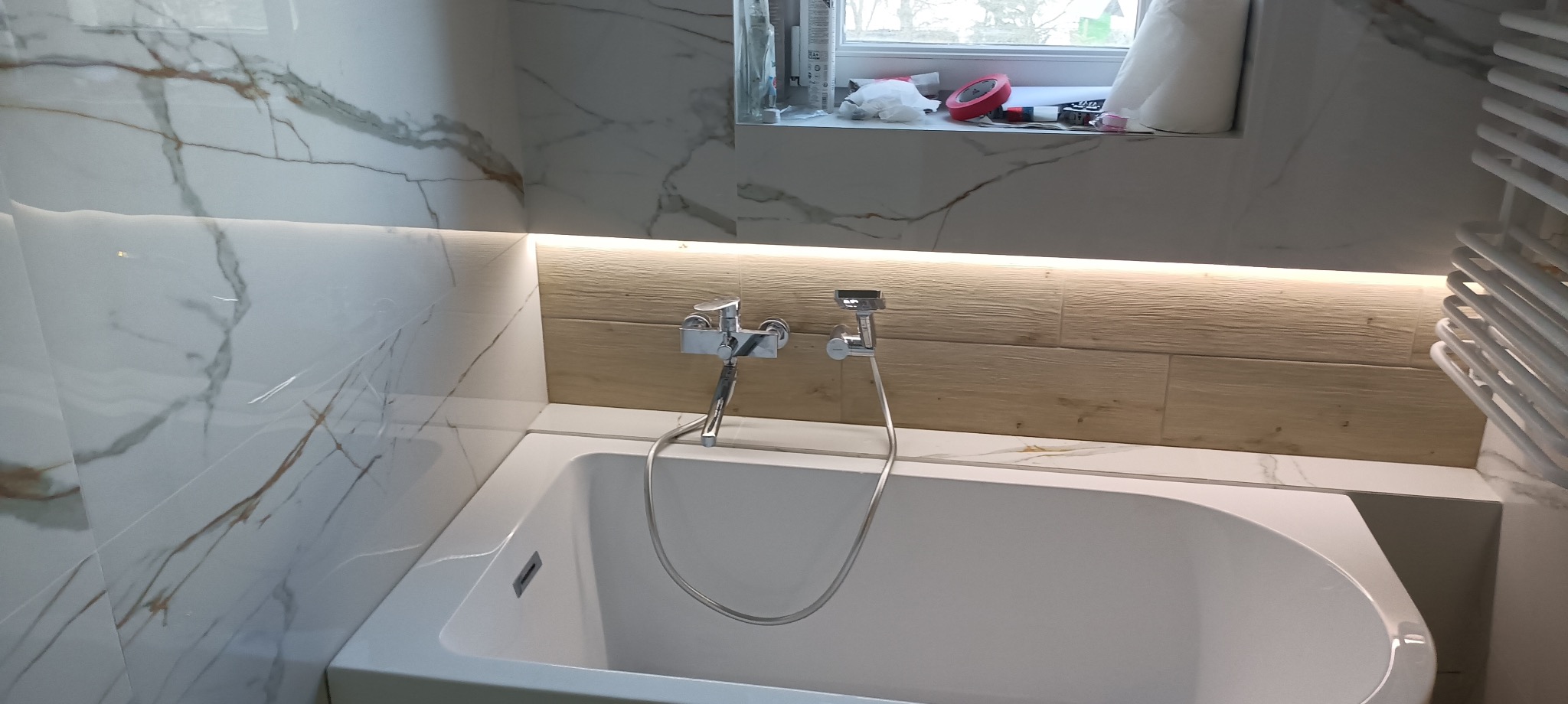 Nowoczesna łazienka z wanną, armaturą i podświetleniem LED. Marmurowe płytki ścienne z wyraźnym żyłowaniem. Drewnopodobne płytki nad wanną. Minimalistyczny design.