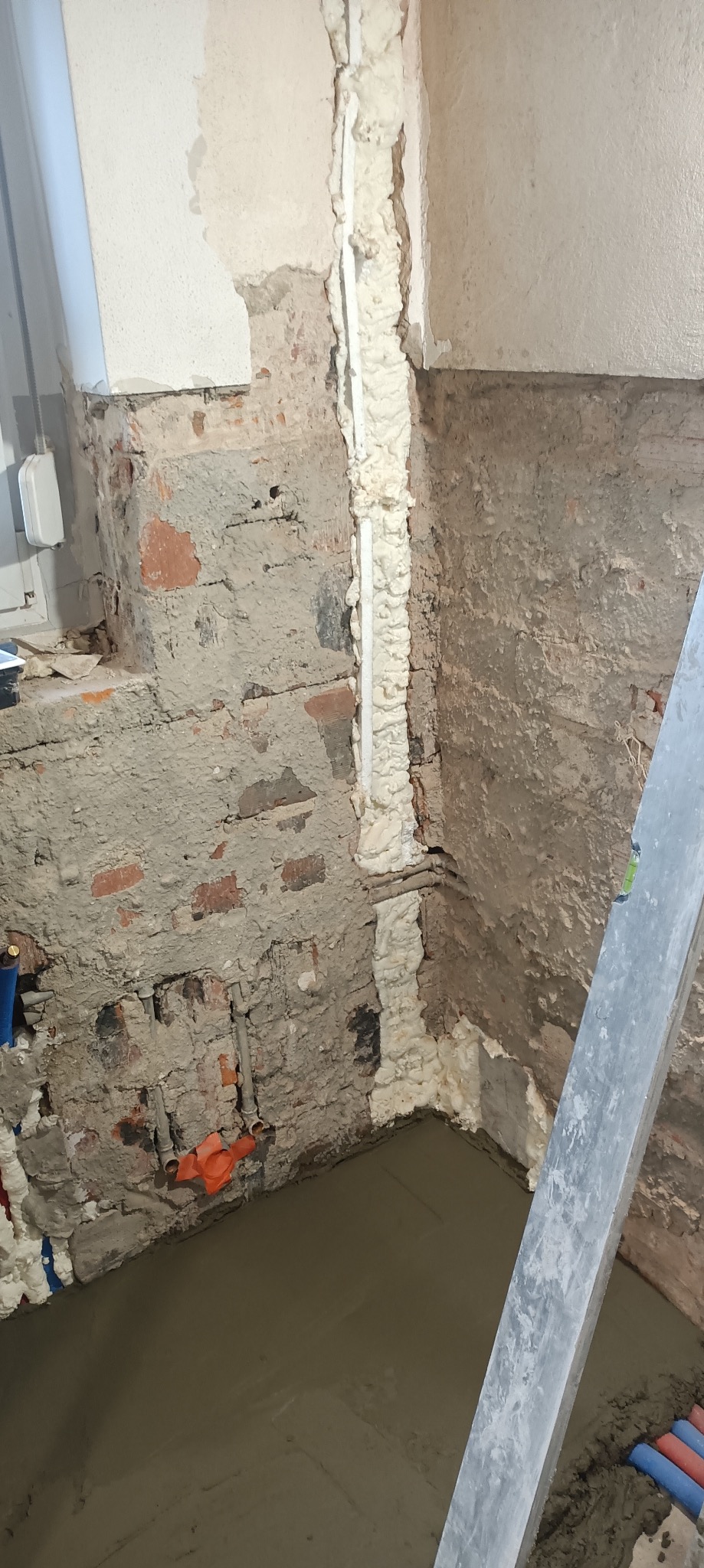 Surowa ściana z instalacją zabezpieczoną pianką montażową i wylewką podłogową w trakcie remontu łazienki. Widoczne rury i fragment drabiny.