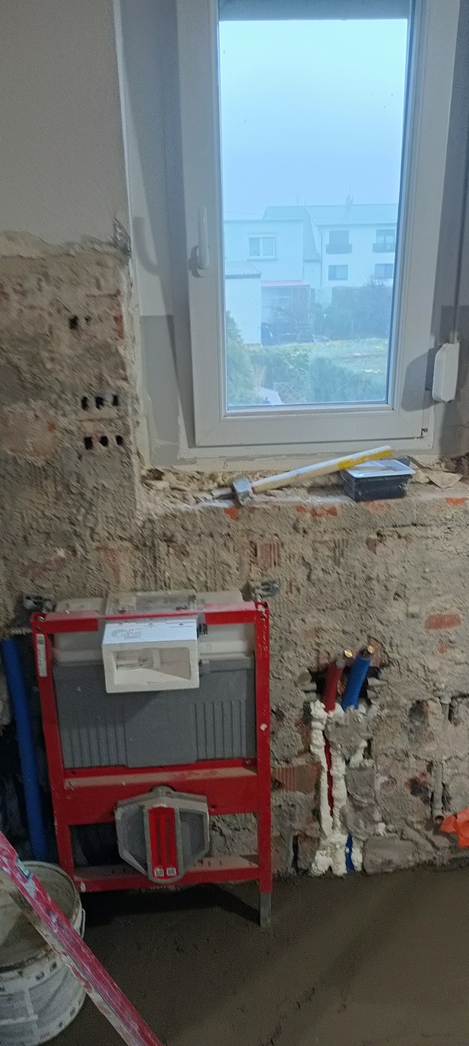 Stelaż podtynkowy WC w trakcie montażu na tle surowej ściany z instalacją wodną i oknem. Widoczne narzędzia i przygotowana podłoga. Etap prac w łazience.