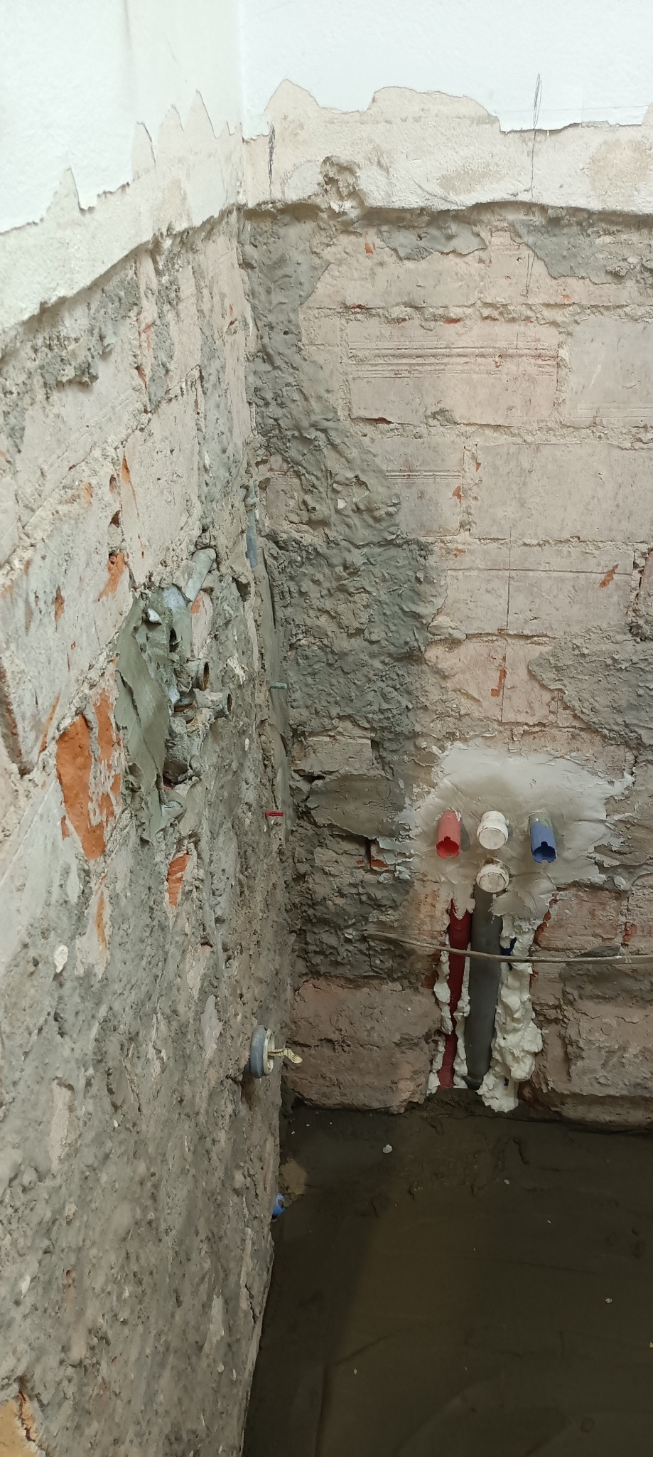 Surowa ściana z cegły i betonu w trakcie remontu łazienki. Widoczne rury wodne z kolorowymi końcówkami i pianka montażowa. Fragment ściany pokryty świeżym tynkiem.