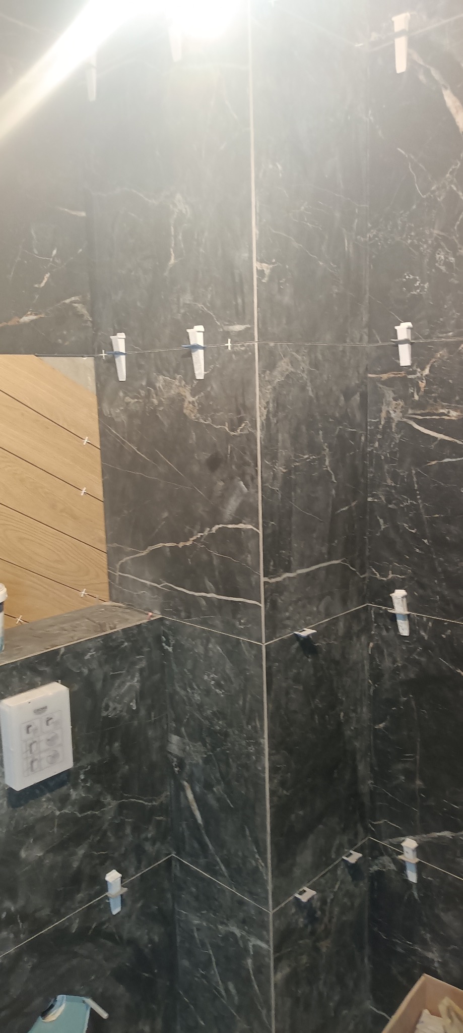 Ściana w trakcie układania płytek imitujących marmur, z użyciem białych krzyżyków dystansowych. Fragment drewnianej zabudowy widoczny po lewej stronie.