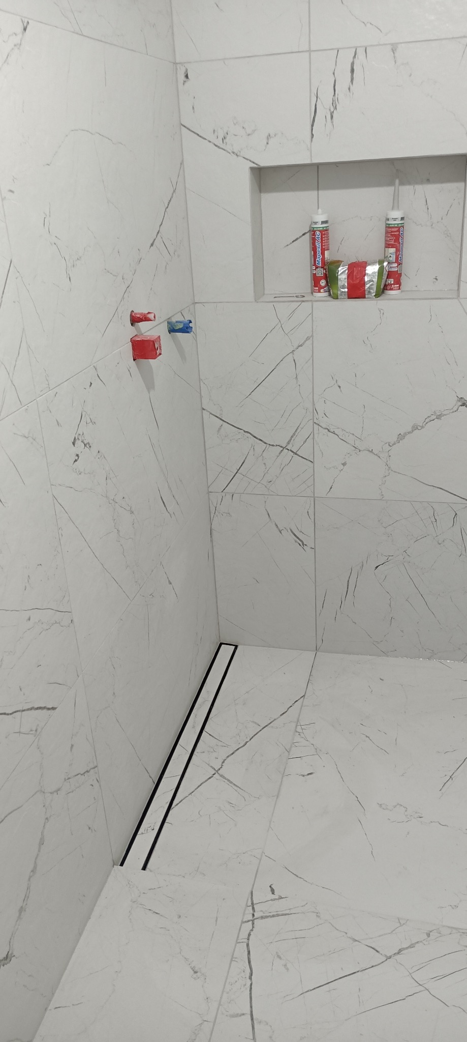 Wykończona łazienka z białymi płytkami imitującymi marmur, widoczny odpływ liniowy, wnęka z materiałami budowlanymi i wystające rury. Minimalistyczny design.