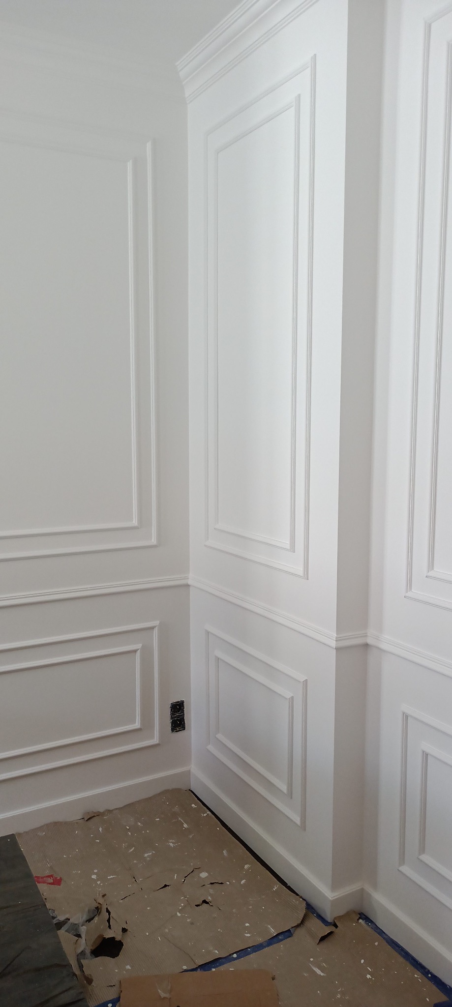 Białe ściany z dekoracyjnymi listwami i sztukaterią w rogu pomieszczenia. Na podłodze karton zabezpieczający. Widoczne gniazdko elektryczne.