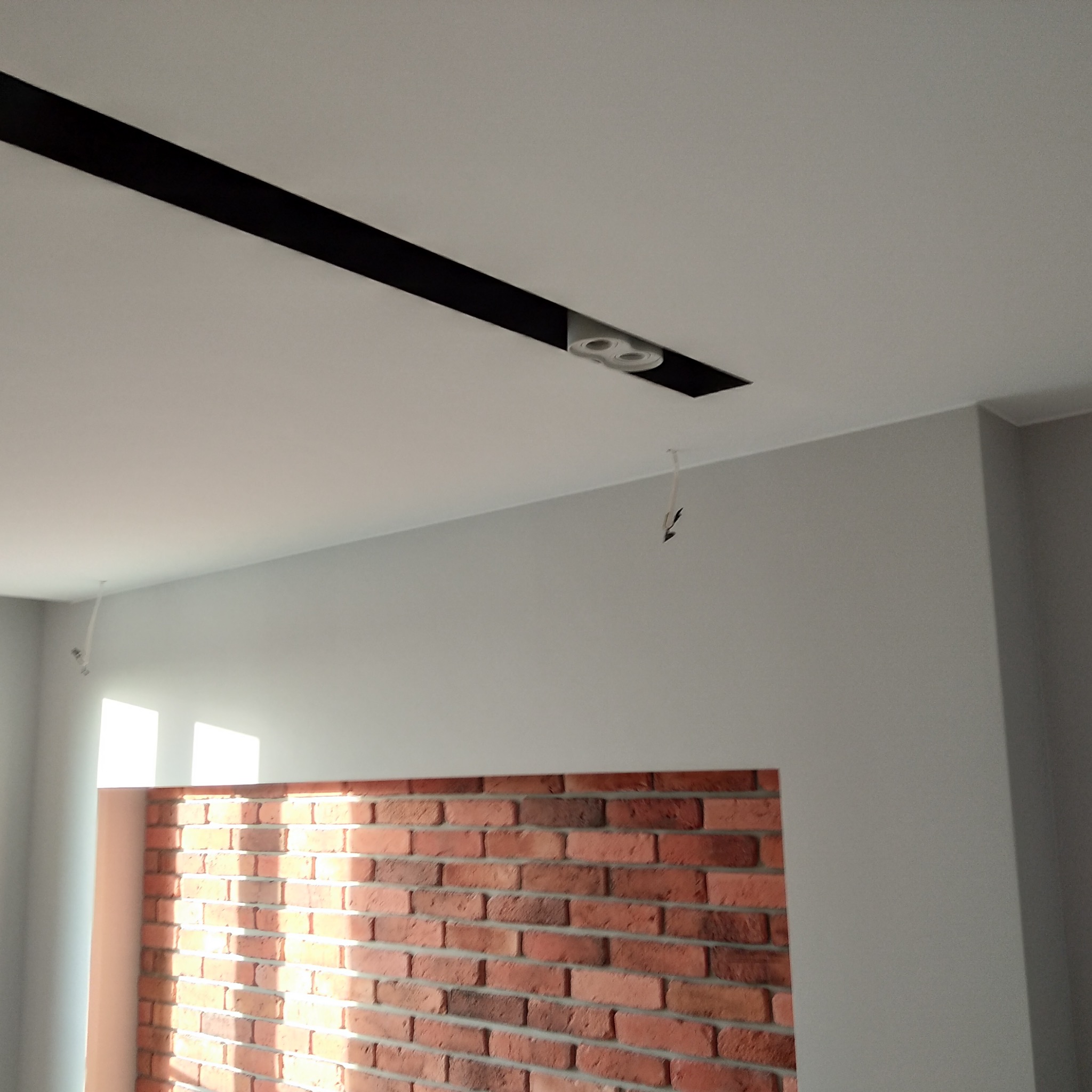 Szara ściana z dekoracyjną czerwoną cegłą i nowoczesnym oświetleniem sufitowym z czarną listwą. Minimalistyczny design wnętrza, widoczne przewody.