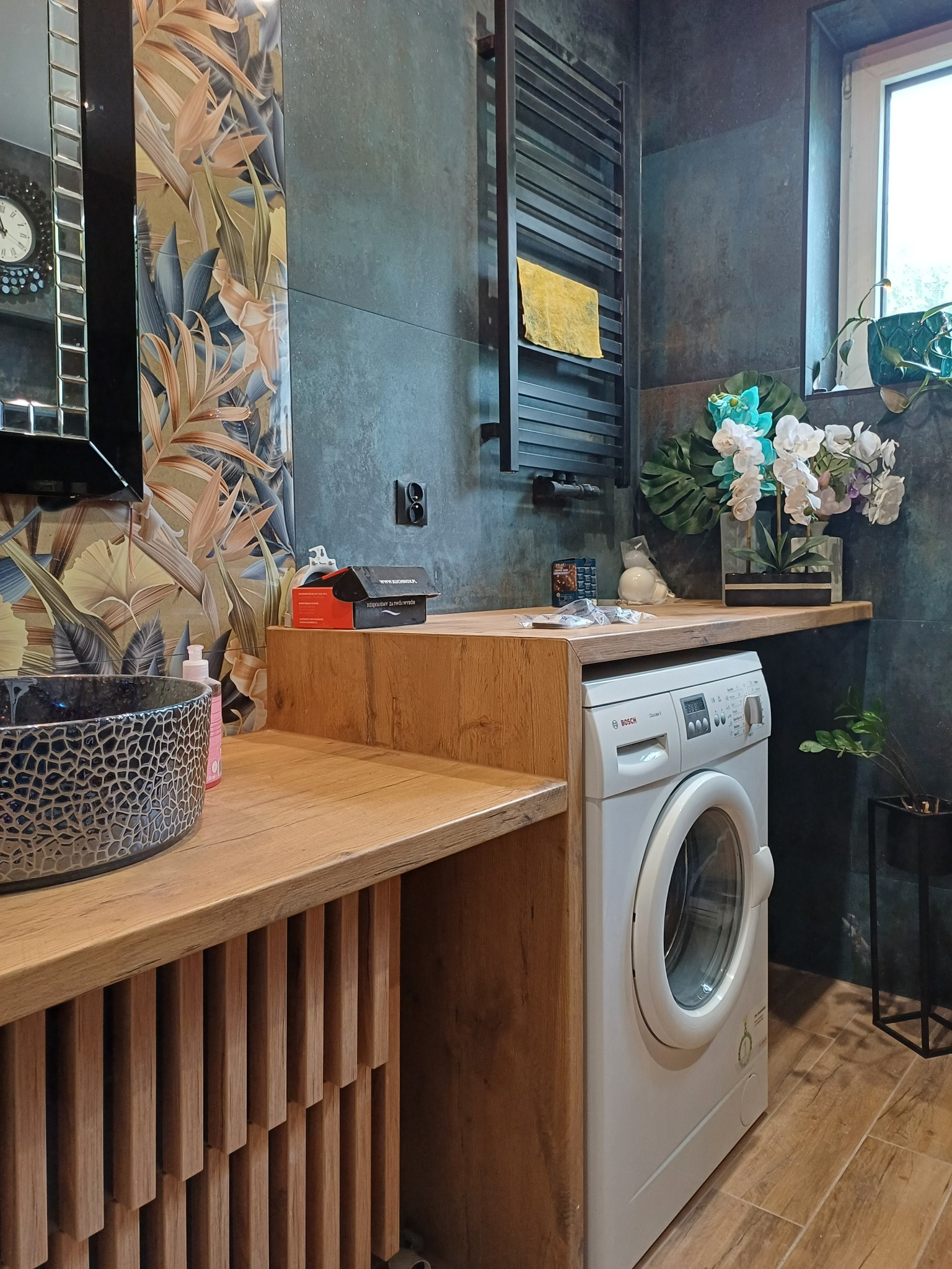 Stylowa łazienka z umywalką w etniczne wzory, drewnianym blatem i pralką Bosch. Na ścianie ciemne płytki i grzejnik drabinkowy. Dekoracje roślinne dodają świeżości.