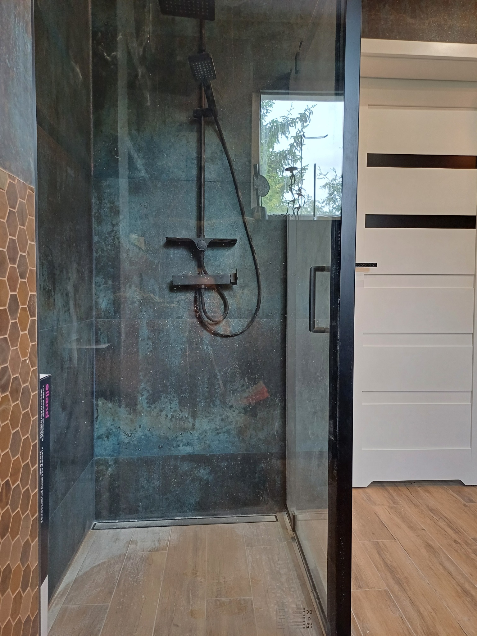 Nowoczesna kabina prysznicowa z ciemnymi, strukturalnymi płytkami i deszczownicą, widok z boku, z elementami dekoracyjnymi i drewnopodobną podłogą.