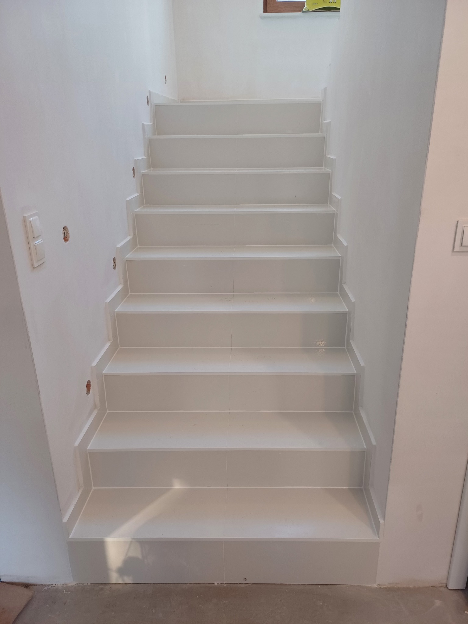 Białe, wykafelkowane schody w jasnym wnętrzu. Widoczne płytki na stopniach i ścianach, z delikatnymi fugami. Minimalistyczny design, surowe wykończenie.