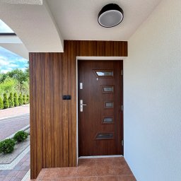 BarTix-Art - Wejście do domu z brązowymi drzwiami i drewnianą boazerią. Biała elewacja z fakturą baranka. Ciemna lampa sufitowa. Brązowa terakota na podłodze. Widok na zieleń.