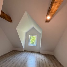 BarTix-Art - Poddasze po remoncie: białe ściany, drewniana podłoga, okno w szczycie. Widoczne drewniane belki stropowe z żarówką. Ukośne ściany i skosy sufitowe.