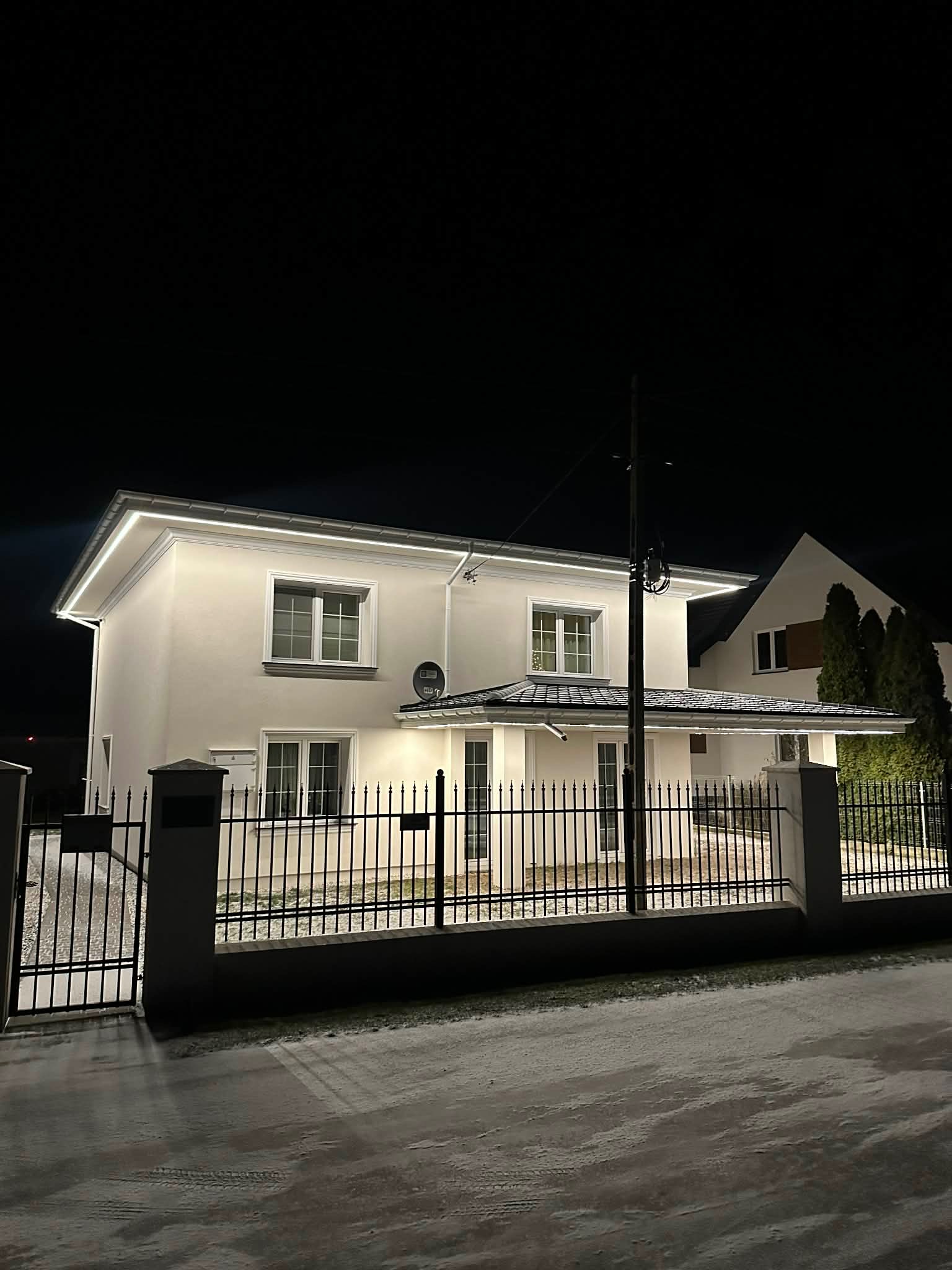 Oświetlona elewacja domu jednorodzinnego nocą, widok zza czarnego, metalowego ogrodzenia. Biała fasada budynku z oknami i podświetleniem LED wzdłuż dachu.