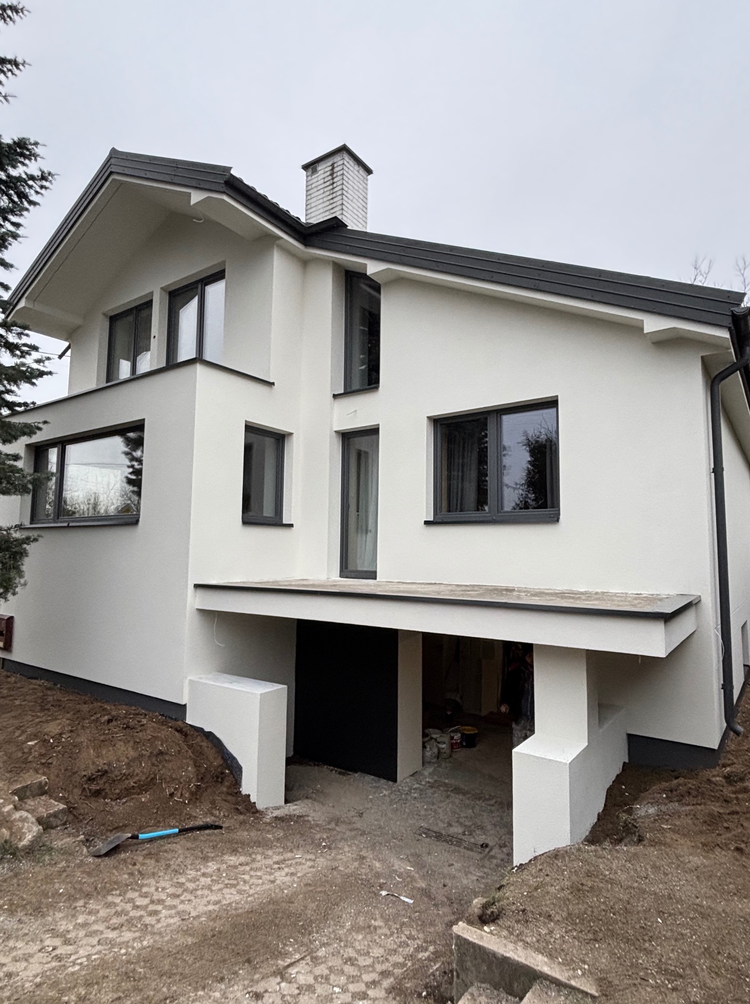 Nowoczesny dom z białymi ścianami i ciemnymi oknami po tynkowaniu. Widoczny fragment ogrodu i narzędzie. Minimalistyczny design, świeżo położony tynk.