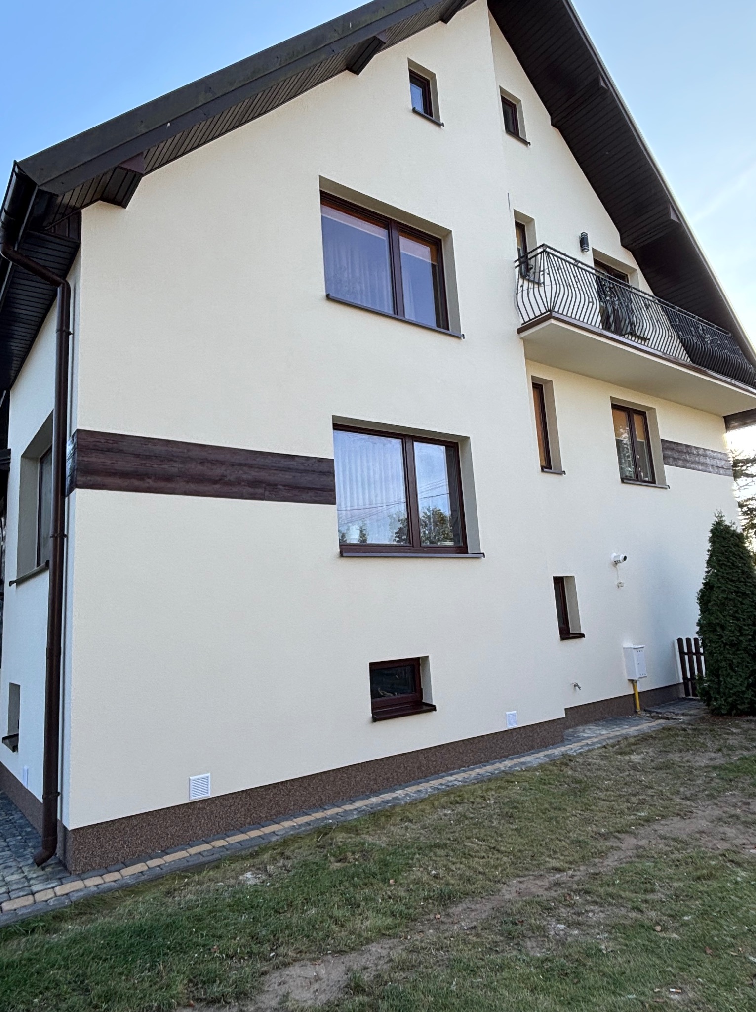 Elewacja domu po renowacji: kremowa ściana z brązowymi akcentami, ciemne okna i balkon z kutą balustradą. Widok z boku, trawnik i kostka brukowa u podstawy.