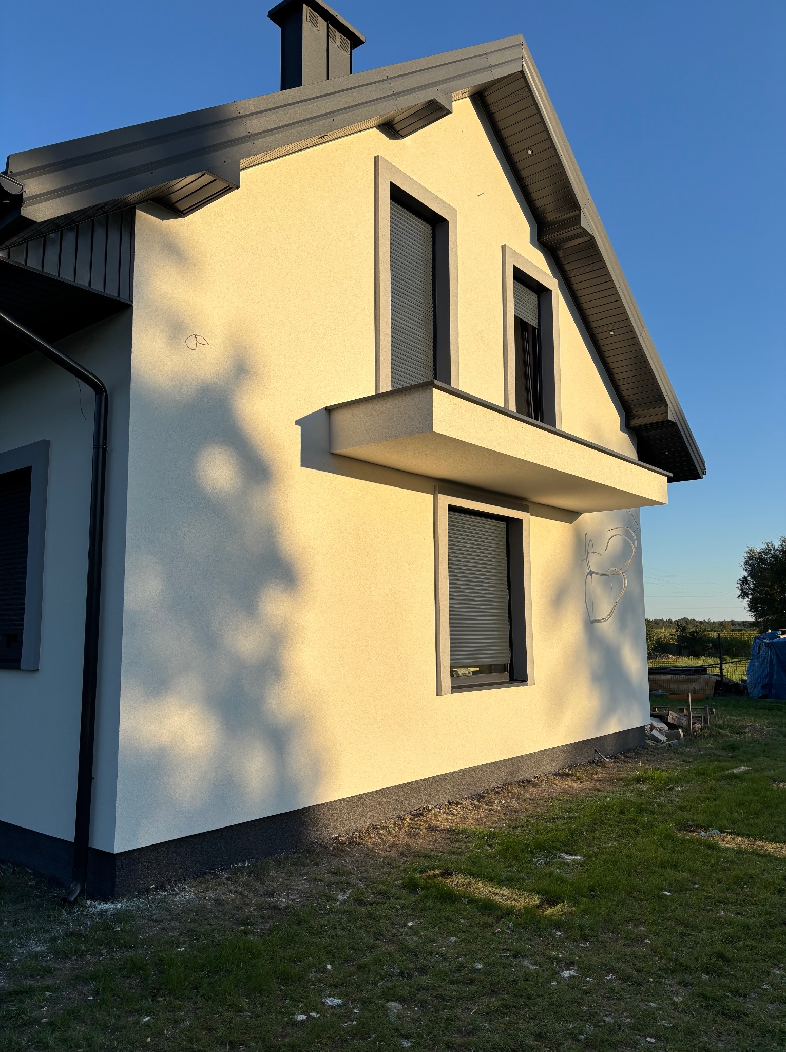 Elewacja domu jednorodzinnego w jasnym odcieniu z szarymi elementami wykończeniowymi, widoczna w popołudniowym słońcu, z fragmentem ogrodu w tle.