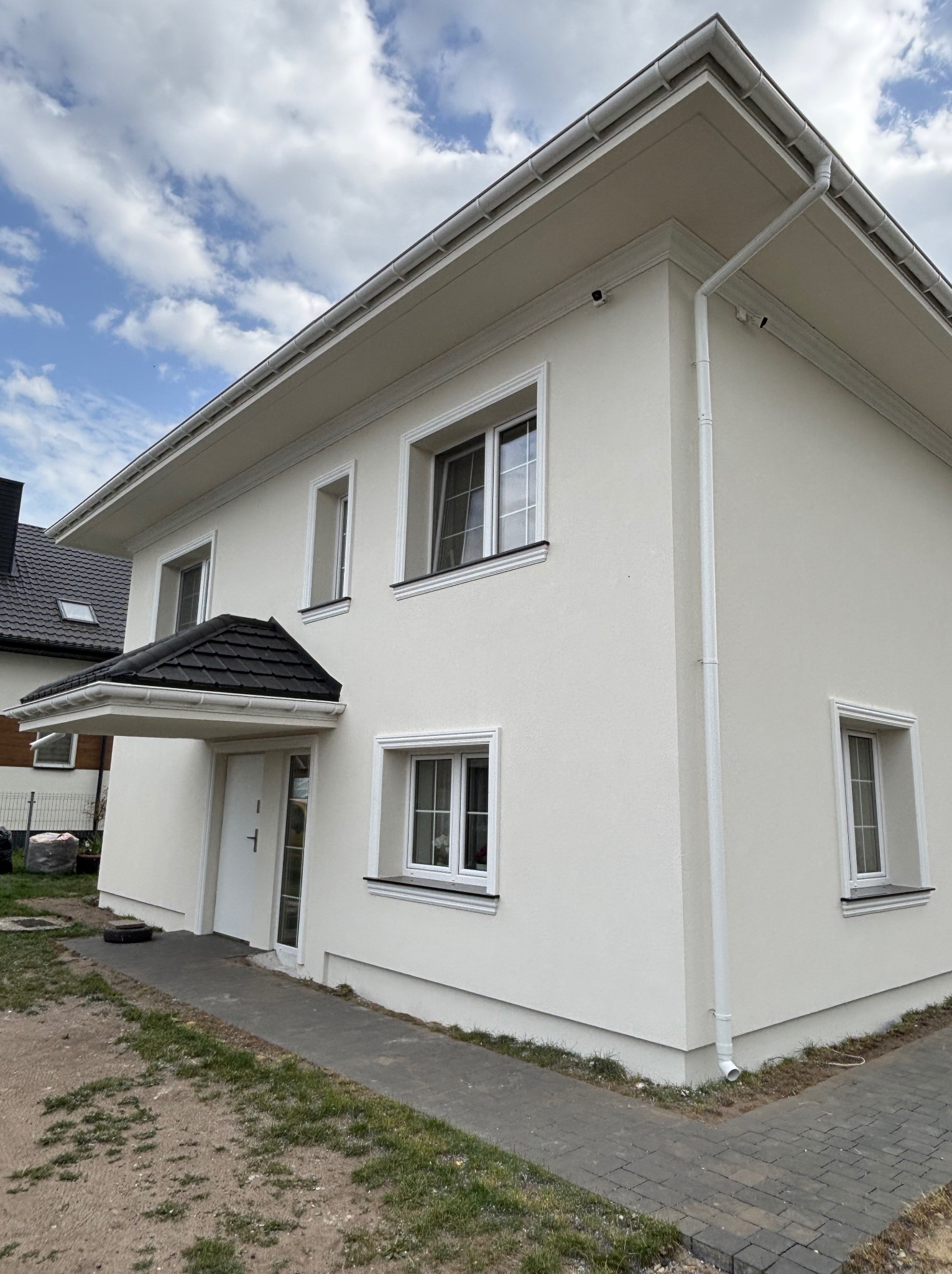 Biały dom jednorodzinny z wykończoną elewacją, widok z dołu, błękitne niebo z chmurami w tle. Detale architektoniczne: białe obramowania okien i rynny.