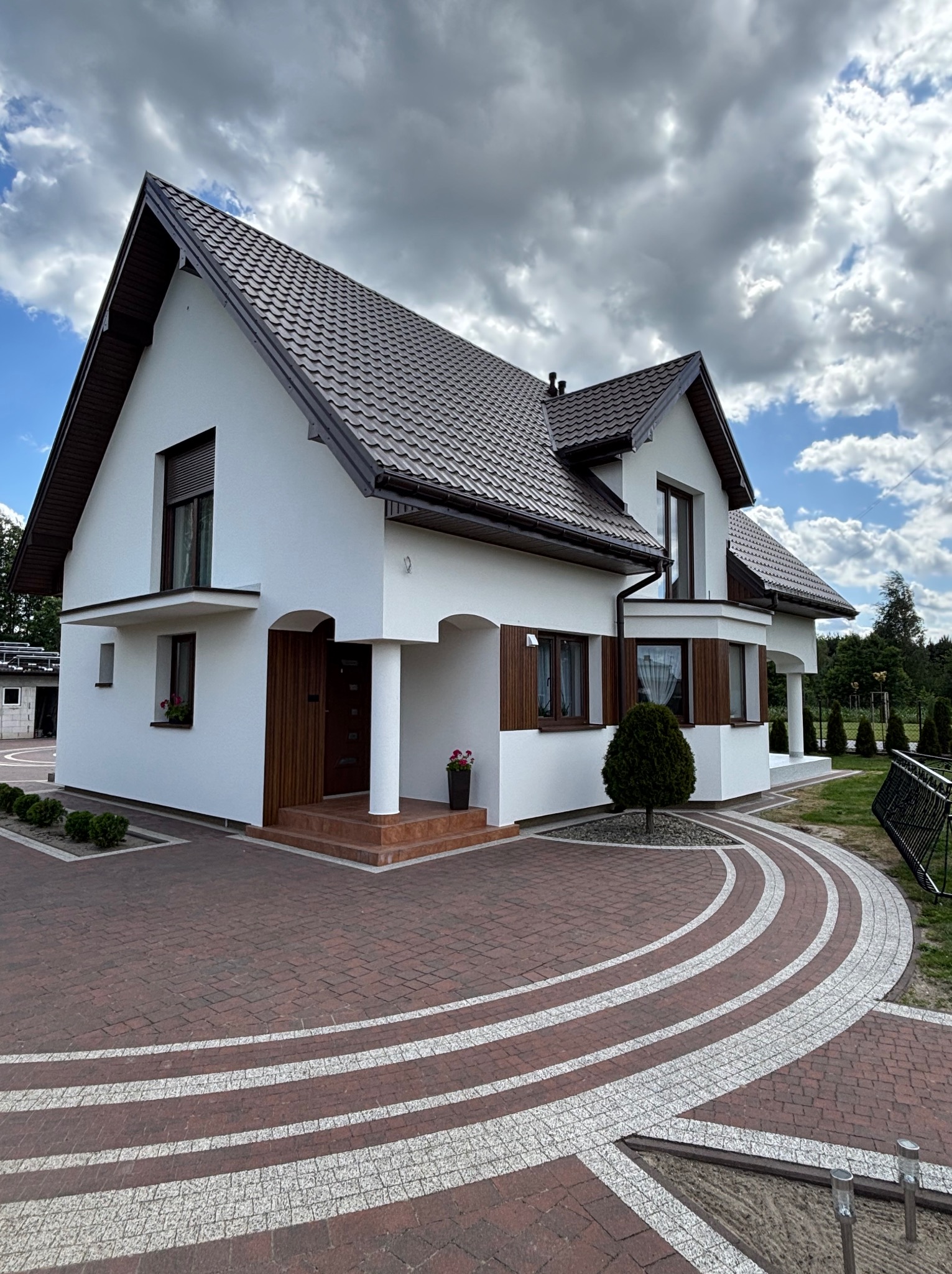 Elewacja domu jednorodzinnego z brązowym dachem i stolarką okienną. Biała fasada, kolumny przy wejściu, dekoracyjny chodnik z kostki. Ujęcie w słoneczny dzień.