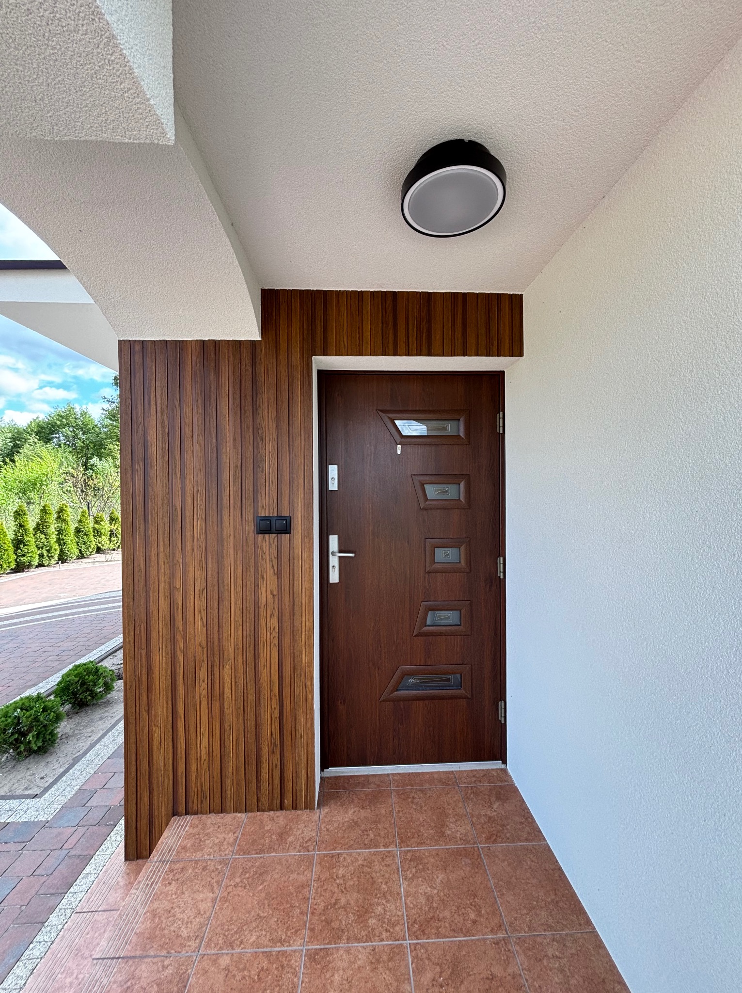 Wejście do domu z brązowymi drzwiami i drewnianą boazerią. Biała elewacja z fakturą baranka. Ciemna lampa sufitowa. Brązowa terakota na podłodze. Widok na zieleń.
