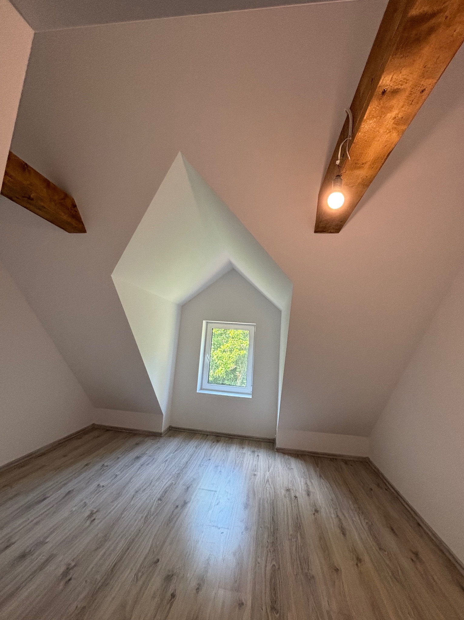 Poddasze po remoncie: białe ściany, drewniana podłoga, okno w szczycie. Widoczne drewniane belki stropowe z żarówką. Ukośne ściany i skosy sufitowe.
