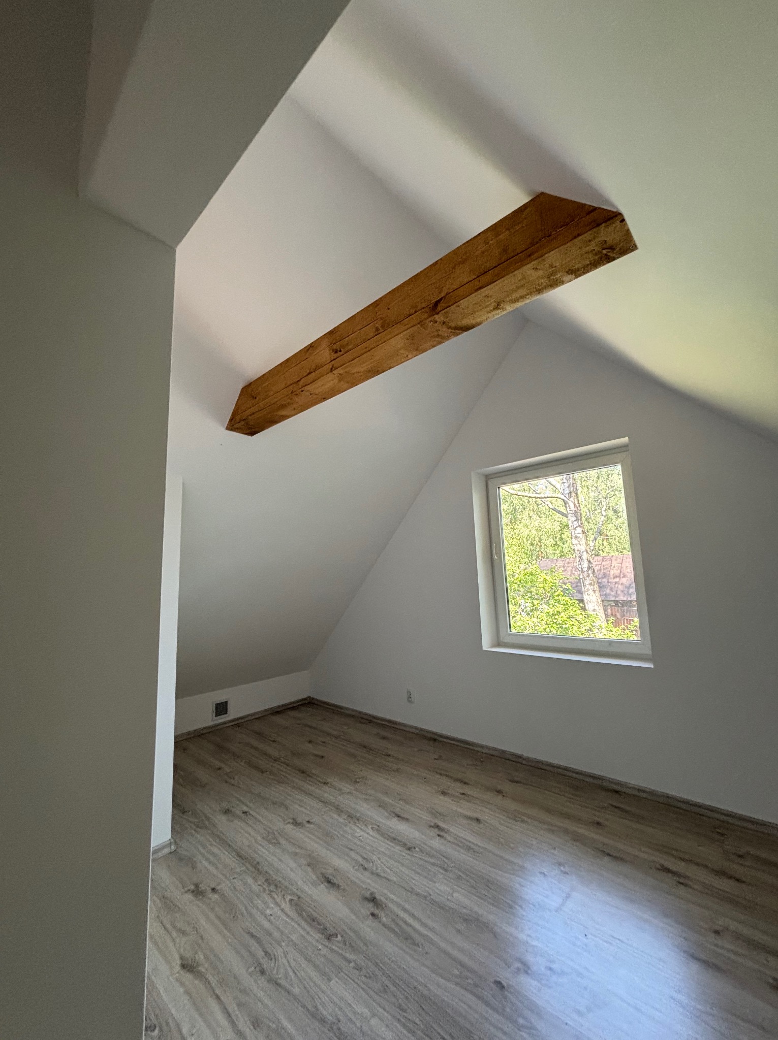 Poddasze po remoncie: drewniana belka, okno z widokiem na zieleń, panele podłogowe imitujące drewno, świeżo pomalowane ściany na biało. Minimalistyczny styl.