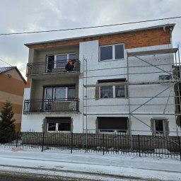 Drew trans bud - Dwupiętrowy budynek w trakcie ocieplania elewacji, widoczne rusztowania i pracownik na balkonie. Część ściany pokryta styropianem, reszta w stanie surowym. Zima, śnieg na ziemi.