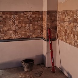 Drew trans bud - Układanie płytek ściennych z wzorem w kuchni podczas remontu. Widoczny poziomica, wiadro i worek z zaprawą. Częściowo wykończona ściana, surowy stan pomieszczenia.