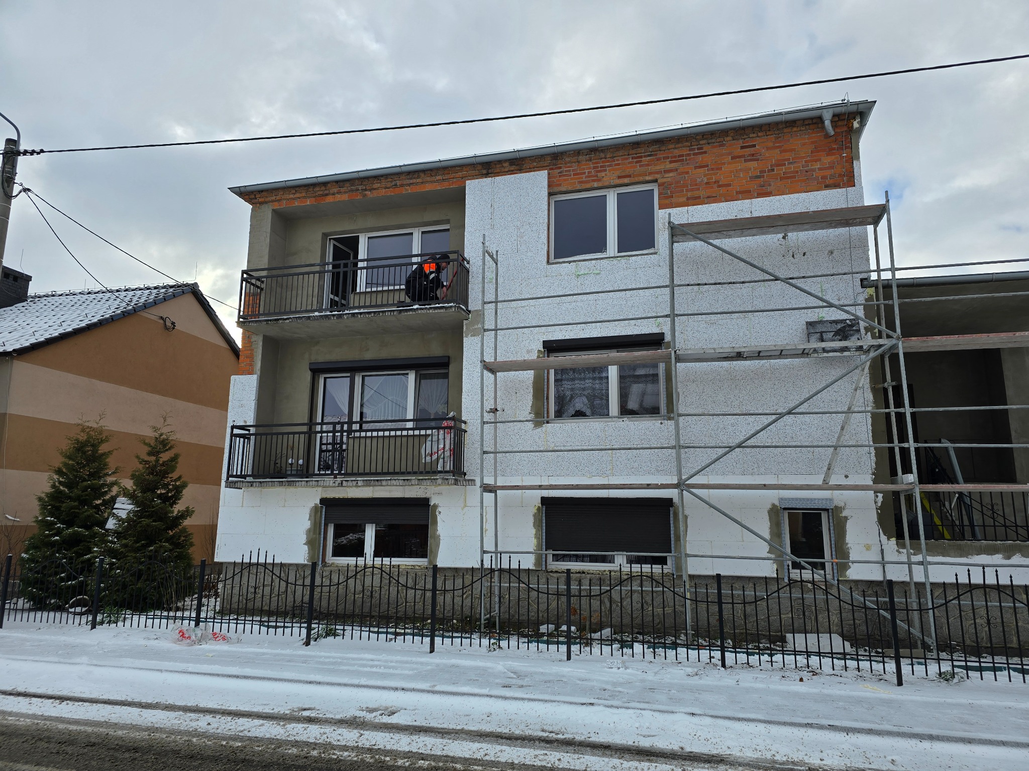 Dwupiętrowy budynek w trakcie ocieplania elewacji, widoczne rusztowania i pracownik na balkonie. Część ściany pokryta styropianem, reszta w stanie surowym. Zima, śnieg na ziemi.