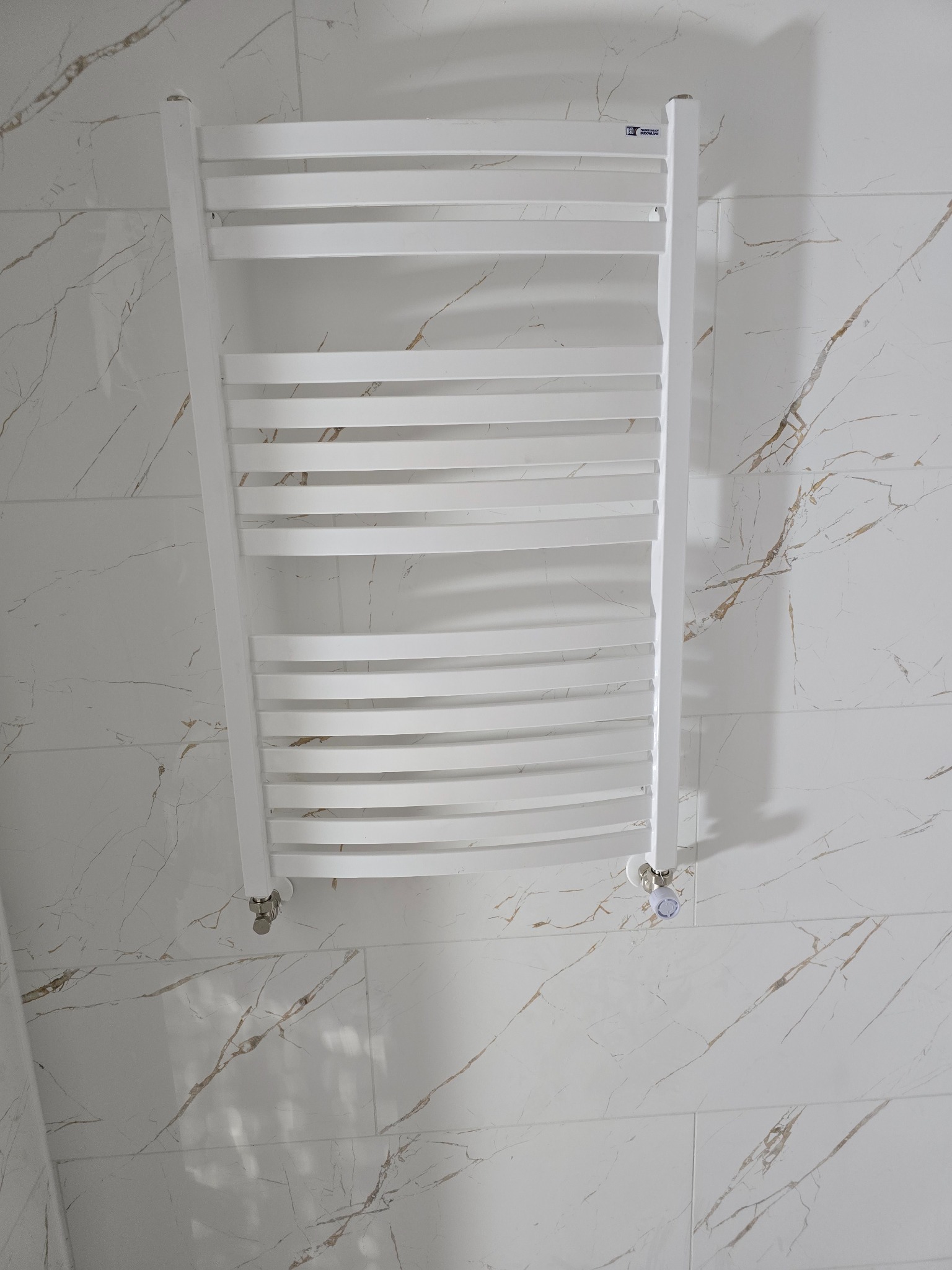 Biały, drabinkowy grzejnik łazienkowy na ścianie z płytek imitujących marmur. Nowoczesny design, widoczne zawory. Ukończony remont łazienki.