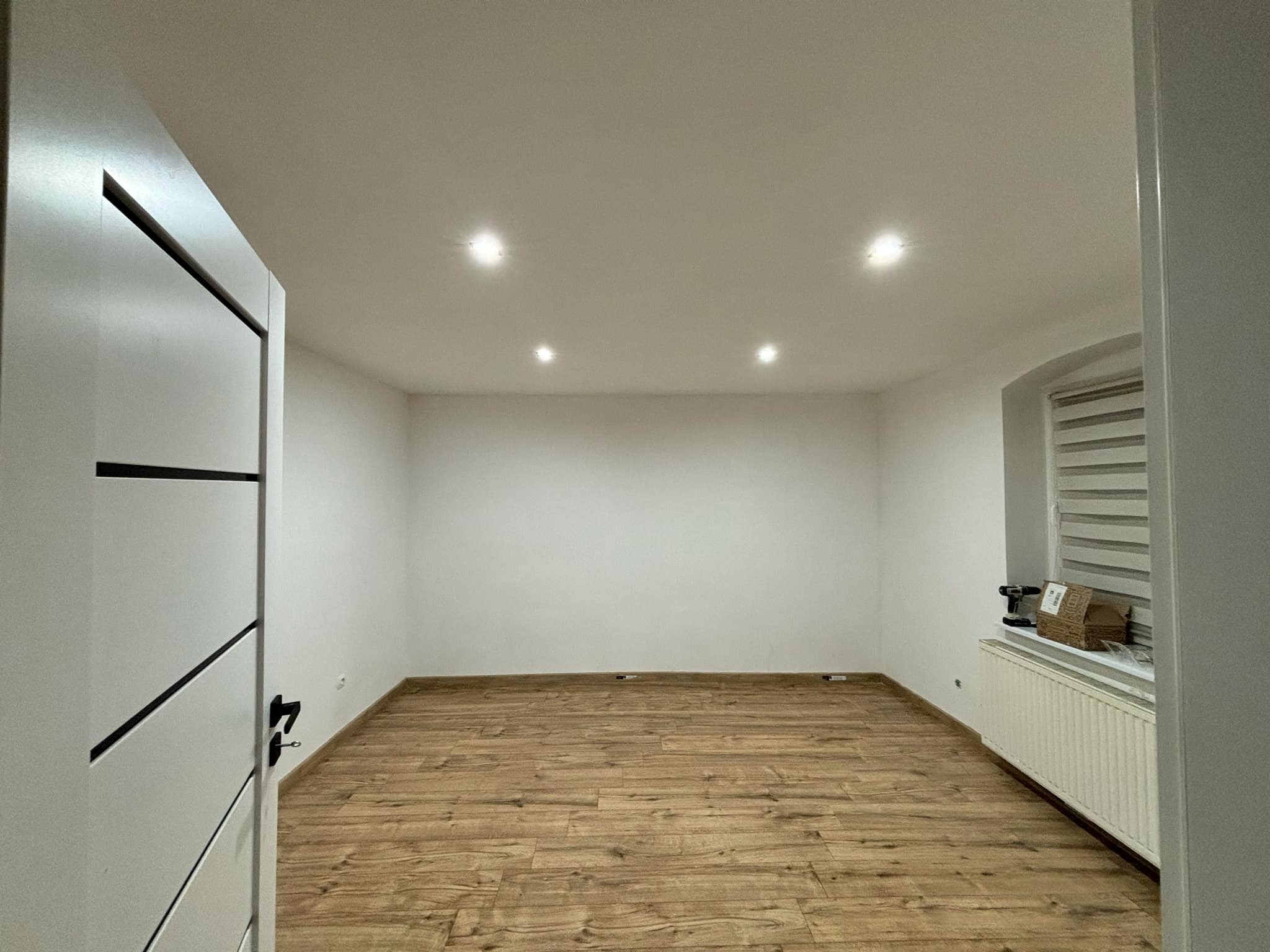 Wykończone wnętrze pokoju z drewnianą podłogą, białymi ścianami i nowoczesnymi drzwiami z czarnymi akcentami. Widoczne oświetlenie sufitowe i grzejnik pod oknem.