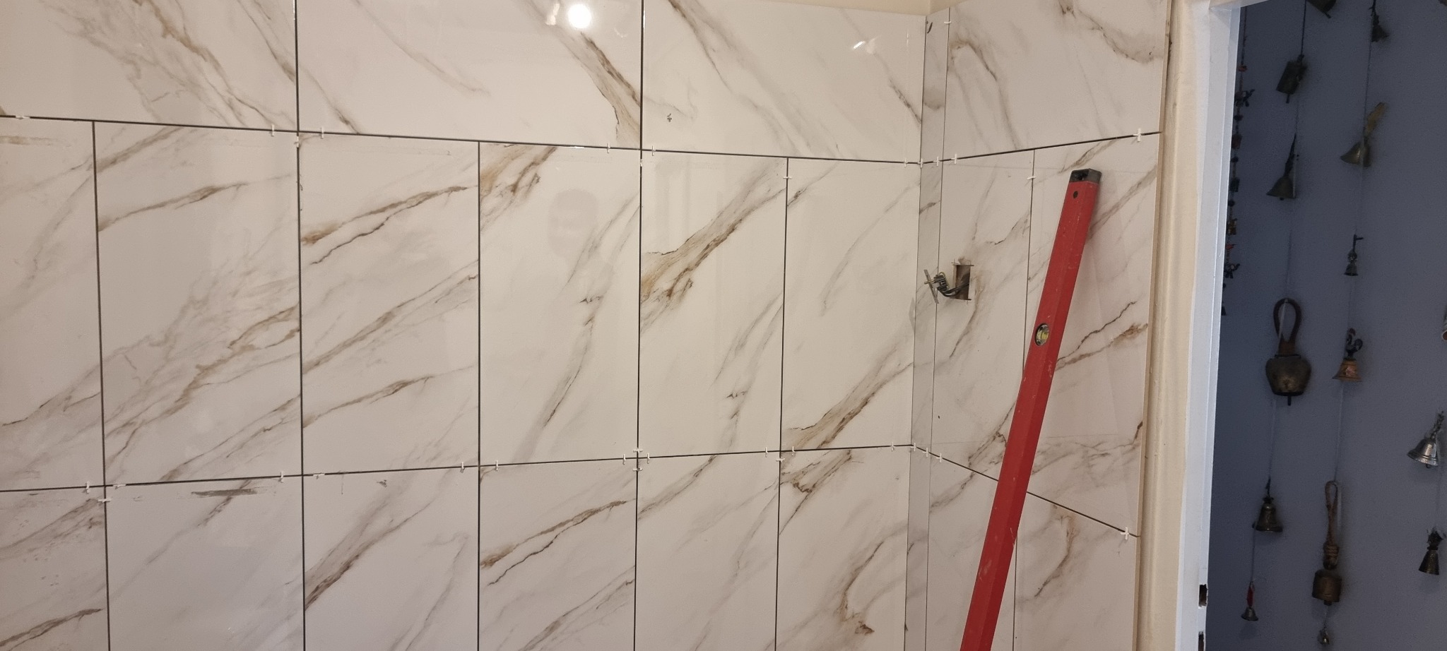 Ściana wyłożona białymi płytkami imitującymi marmur z brązowymi żyłami, poziomica oparta o ścianę, widoczne krzyżyki dystansowe, fragment dekoracji w tle.