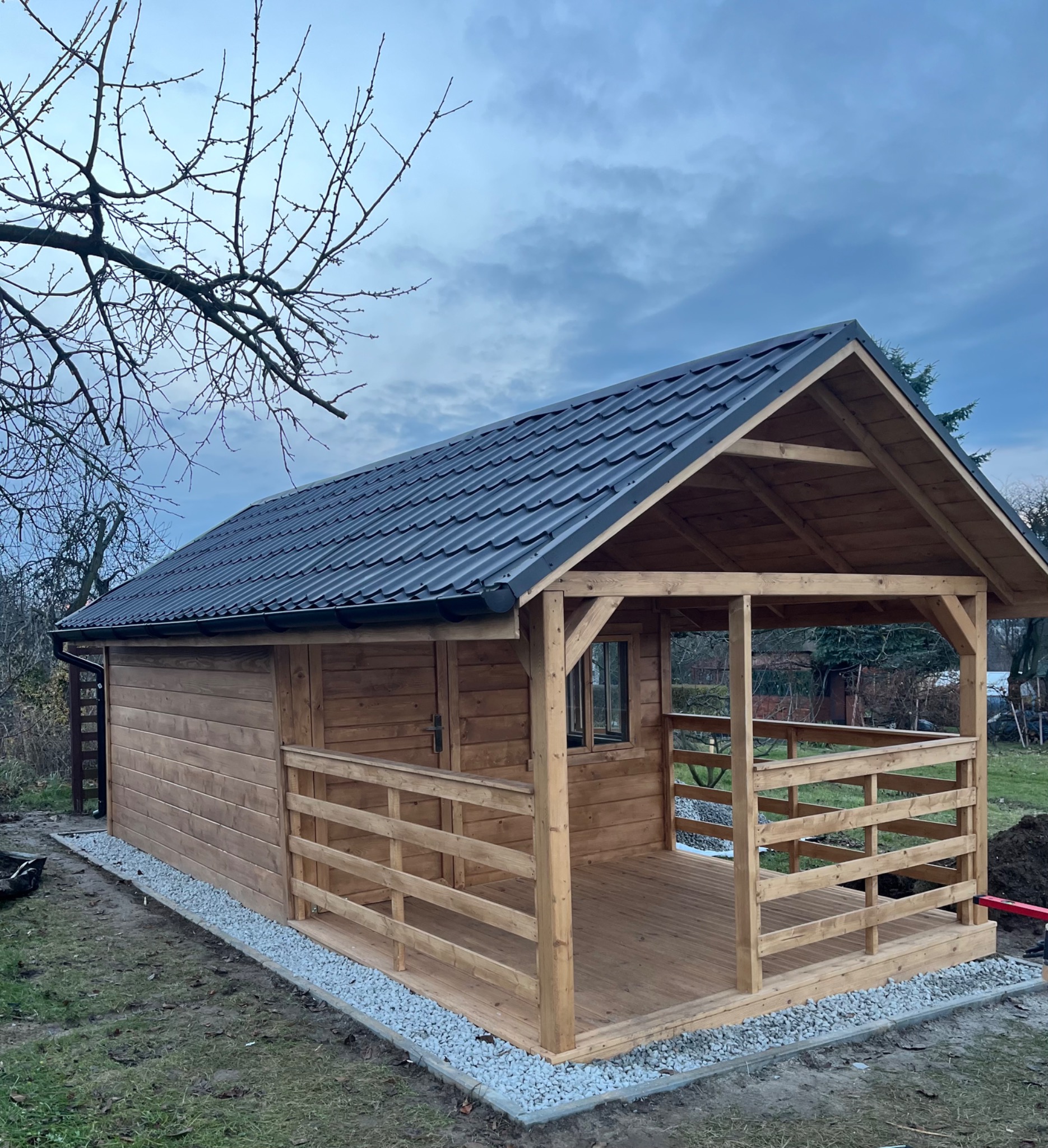 Drewniana altana z zadaszeniem z blachy, otwartą przestrzenią tarasową i balustradami, otoczona zielenią i kamiennym obrzeżem. Widok z boku, pochmurne niebo w tle.