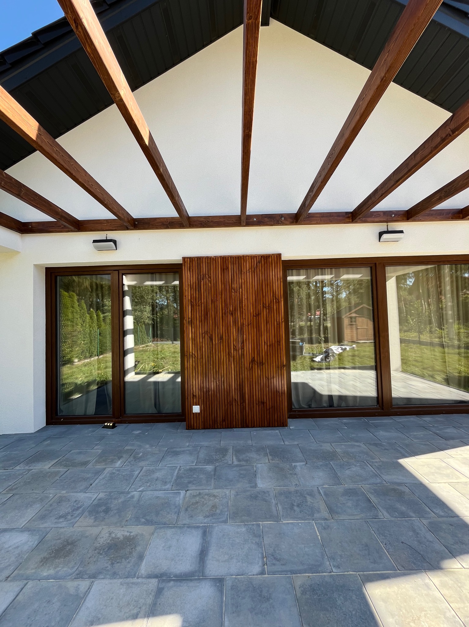 Nowoczesna, drewniana pergola zadaszająca taras z szarych płyt. Widoczne przesuwne drzwi tarasowe i drewniana ścianka działowa. Oświetlenie zewnętrzne.