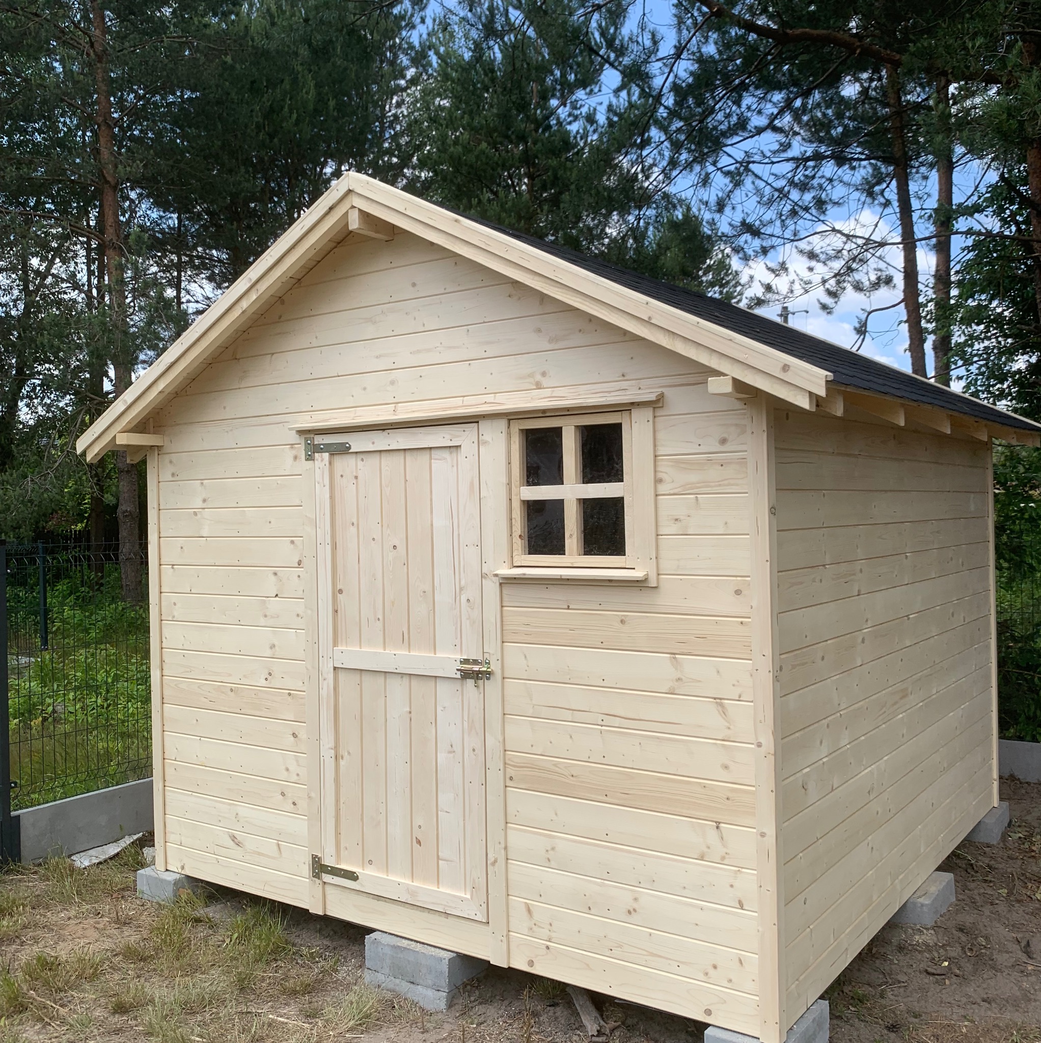 Drewniana konstrukcja z oknem i drzwiami, prawdopodobnie domek narzędziowy, na betonowych bloczkach, otoczona zielenią i drzewami. Widok z przodu, jasne drewno.